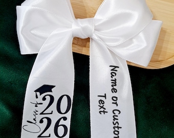 Puede incluir: Un lazo de graduación de satén blanco con largas colas. Una cola dice "Clase de 2026" con un gorro de graduación. La otra cola dice "Nombre o texto personalizado". El lazo está sobre una bandeja de madera.
