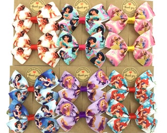 Puede incluir: Nueve pares de lazos para el pelo hechos a mano con diferentes princesas Disney. Cada lazo está hecho con dos capas de tela y tiene una cinta roja, amarilla o dorada en el centro. Los lazos se muestran en cartulina marrón con el texto "Gracias" impreso en cada tarjeta.