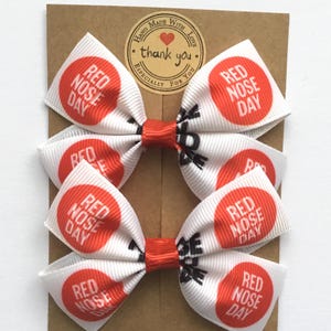 Pair of white grosgrain ribbon hair bows with "Red Nose Day" red circle print. Handmade 3-inch bows for Comic Relief 2026.