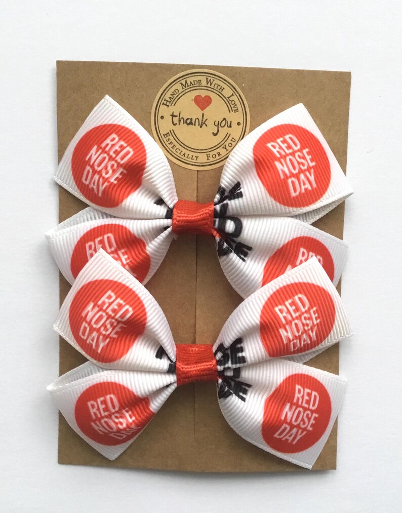 Pair of white grosgrain ribbon hair bows with "Red Nose Day" red circle print. Handmade 3-inch bows for Comic Relief 2026.
