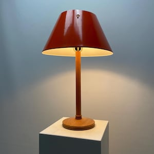 May include: A red lampshade with a wooden base sits on a white surface. The lampshade is lit and casts a warm glow.