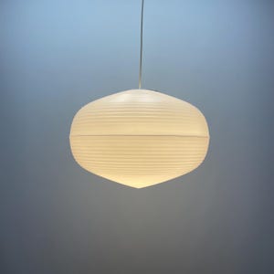 Puede incluir: Una l&aacute;mpara colgante blanca de forma ovalada con textura acanalada. Est&aacute; suspendida de un fino cable blanco. La luz est&aacute; encendida, emitiendo un brillo suave. El fondo es un degradado de azul y gris.