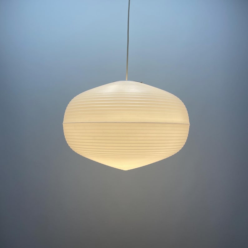 Puede incluir: Una l&aacute;mpara colgante blanca de forma ovalada con textura acanalada. Est&aacute; suspendida de un fino cable blanco. La luz est&aacute; encendida, emitiendo un brillo suave. El fondo es un degradado de azul y gris.