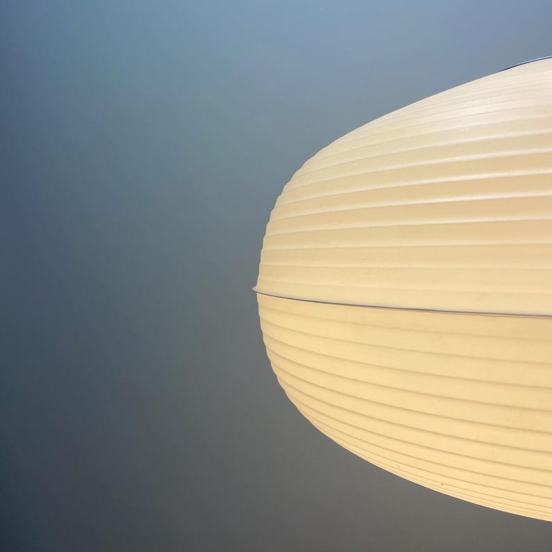 Puede incluir: Primer plano de una l&aacute;mpara colgante ovalada, de color crema y acanalada. La luz est&aacute; encendida y la l&aacute;mpara est&aacute; suspendida por un cable fino. El fondo es un degradado de azul y gris.
