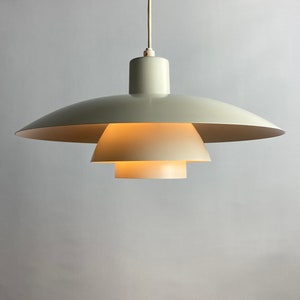 May include: A pale gray pendant light fixture featuring a tiered design with three concentric, bowl-shaped shades of varying sizes. The light is suspended by a single white cord and emits a soft, warm glow from the center of the layered structure.