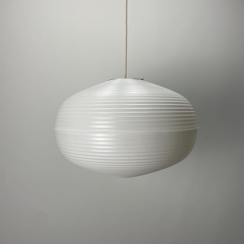Puede incluir: Una l&aacute;mpara colgante esf&eacute;rica blanca con detalles acanalados horizontales. La luz cuelga de un fino cable blanquecino sobre un fondo gris neutro. La l&aacute;mpara tiene un dise&ntilde;o moderno y minimalista.