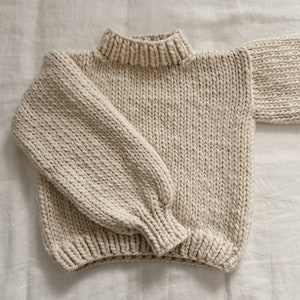 Puede incluir: Un suéter de punto de color crema con cuello alto y mangas largas. El suéter está hecho de un hilo grueso y tiene una textura de punto grueso.