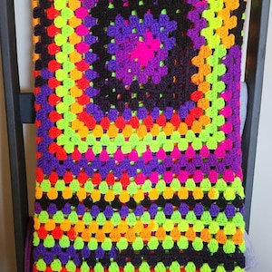 Puede incluir: Una manta de ganchillo colorida con un patrón negro, morado, naranja, verde y rosa. La manta está hecha de cuadrados de la abuela y tiene un diseño circular en el centro.