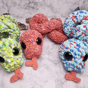 May include: A selection of handmade, plush alien-like creatures in various colours. The creatures have large, black eyes and small, orange feet. The plushies are made of a soft, textured yarn in shades of green, red, blue, and orange.