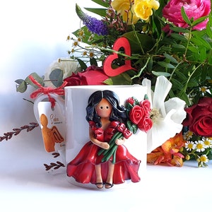 Könnte beinhalten: Weiße Keramiktasse mit einer modellierten Figur einer Frau in einem roten Kleid, die rote Rosen hält. Die Tasse hat ein Etikett mit einer Engel-Illustration. Ein bunter Blumenstrauß, darunter Rosen, befindet sich im Hintergrund.