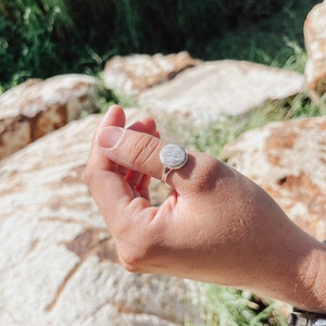 Puede incluir: Un anillo de plata con una piedra redonda, texturizada y gris. El anillo está en el dedo de una persona.