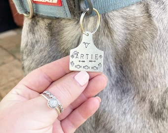 Op de afbeelding: Een zilveren hondenpenning met de naam "ARTIE" erop gegraveerd. De penning heeft een cowboyhoed, cactus en pijlmotieven. De penning is bevestigd aan een blauwe hondenriem.