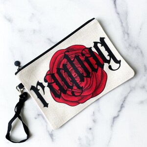 May include: A rectangular canvas pouch with a black zipper and wrist strap. The pouch features a large red rose with the word "FRIEND" in black gothic lettering. The background is a natural off-white color.