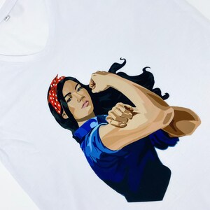 May include: White t-shirt with a graphic of a woman with dark hair and a red bandana, flexing her bicep. The woman is wearing a blue shirt.