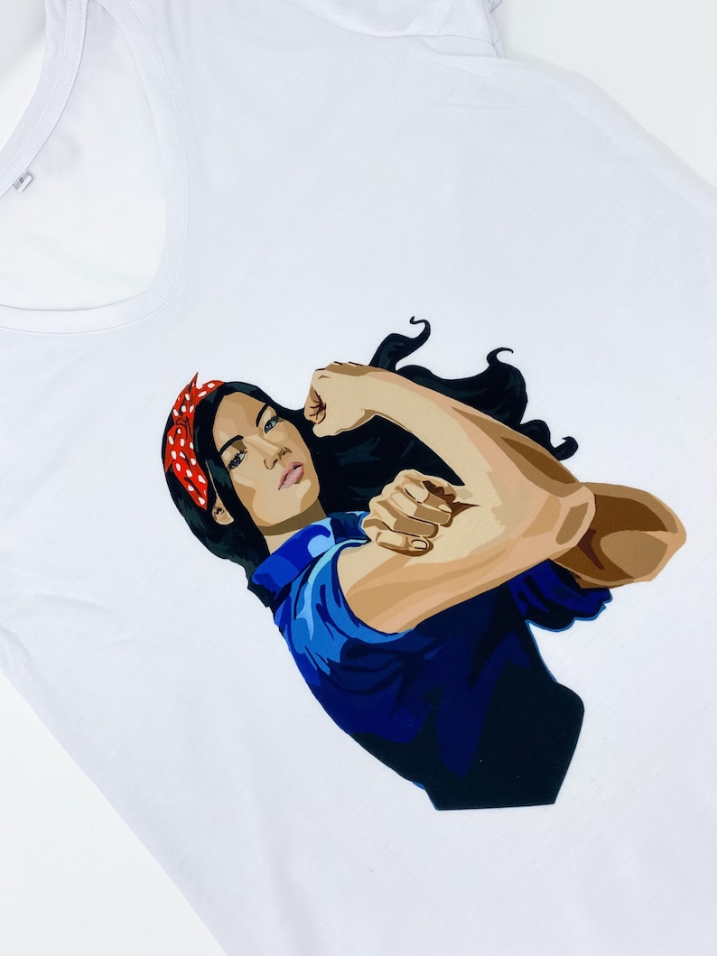 May include: White t-shirt with a graphic of a woman with dark hair and a red bandana, flexing her bicep. The woman is wearing a blue shirt.
