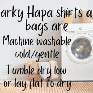 May include: A white washing machine with a white door and a silver knob. The washing machine is in a laundry room with a white wall and a brown rug. The text on the wall reads "Snarky Hapa shirts and bags are Machine washable cold/gentle Tumble dry low or lay flat to dry".