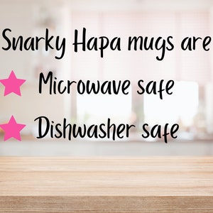 May include: A close-up of a wooden table with a blurred background of a kitchen. The text "Snarky Hapa mugs are" is written in black cursive font. Two pink stars are next to the text "Microwave safe" and "Dishwasher safe".