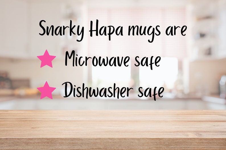 May include: A close-up of a wooden table with a blurred background of a kitchen. The text "Snarky Hapa mugs are" is written in black cursive font. Two pink stars are next to the text "Microwave safe" and "Dishwasher safe".