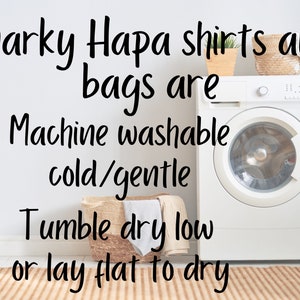 May include: A white washing machine with a white door and a silver knob. The washing machine is in a laundry room with a white wall and a brown rug. The text on the wall reads "Snarky Hapa shirts and bags are Machine washable cold/gentle Tumble dry low or lay flat to dry".