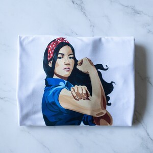 May include: White t-shirt with a graphic of a woman with a red and white polka dot headband, blue shirt, and a flexed arm.