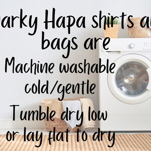 May include: A white washing machine with a white door and a silver knob. The text "Snarky Hapa shirts and bags are Machine washable cold/gentle Tumble dry low or lay flat to dry" is written on a white wall behind the washing machine.