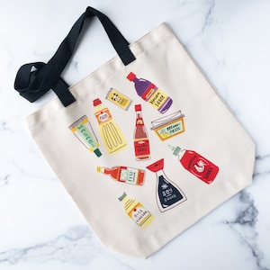 May include: A white canvas tote bag with black handles features a colorful illustration of various Asian cooking ingredients, including soy sauce, fish sauce, rice vinegar, miso paste, and sriracha sauce.