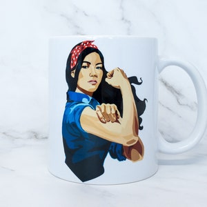 May include: White ceramic mug featuring a graphic of a woman flexing her arm, wearing a red polka dot bandana and a blue collared shirt. The mug has a white handle and a white interior.