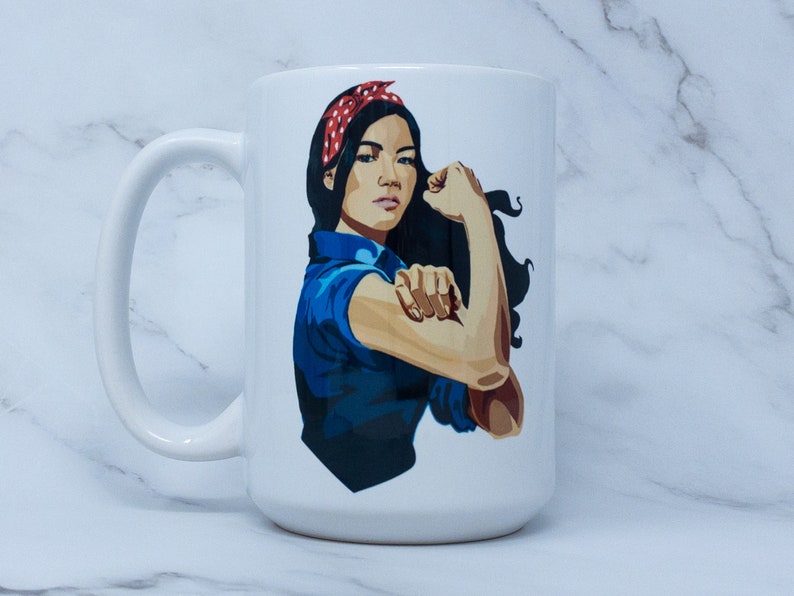 May include: White ceramic mug with a colorful illustration of a woman with her arm raised in a 'we can do it' pose. She is wearing a blue shirt and a red and white polka dot headband.