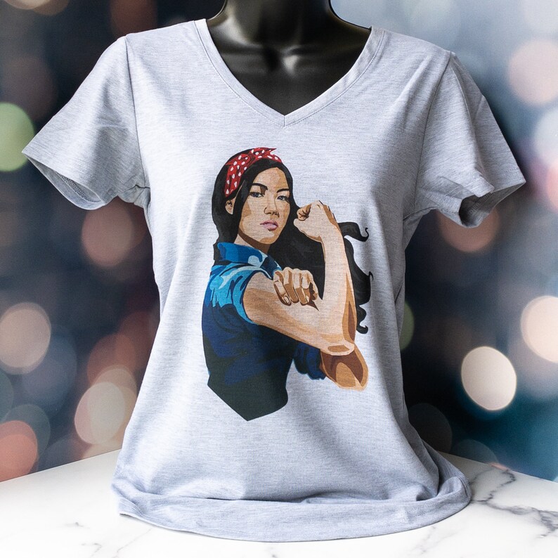 May include: Gray V-neck t-shirt with a graphic print of a woman with a red headband, flexing her bicep. The woman is wearing a blue shirt and has dark hair.