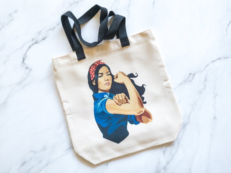 May include: A white canvas tote bag with black handles features a colorful illustration of a woman with her arm raised in a powerful pose. She is wearing a blue shirt, a red and white polka dot bandana, and has long dark hair.