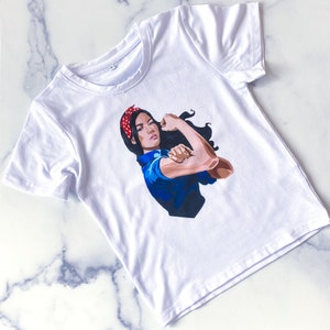 May include: White t-shirt featuring a graphic of a woman flexing her arm, inspired by the Rosie the Riveter image. The woman has black hair, a red polka-dot headband, and is wearing a blue collared shirt. The graphic is centered on the front of the tee.