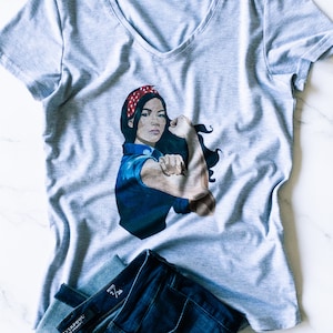 May include: A gray V-neck t-shirt with a graphic of a woman with a red bandana and a blue shirt, flexing her bicep. The woman is depicted in a classic "We Can Do It" pose. The t-shirt is paired with blue jeans.