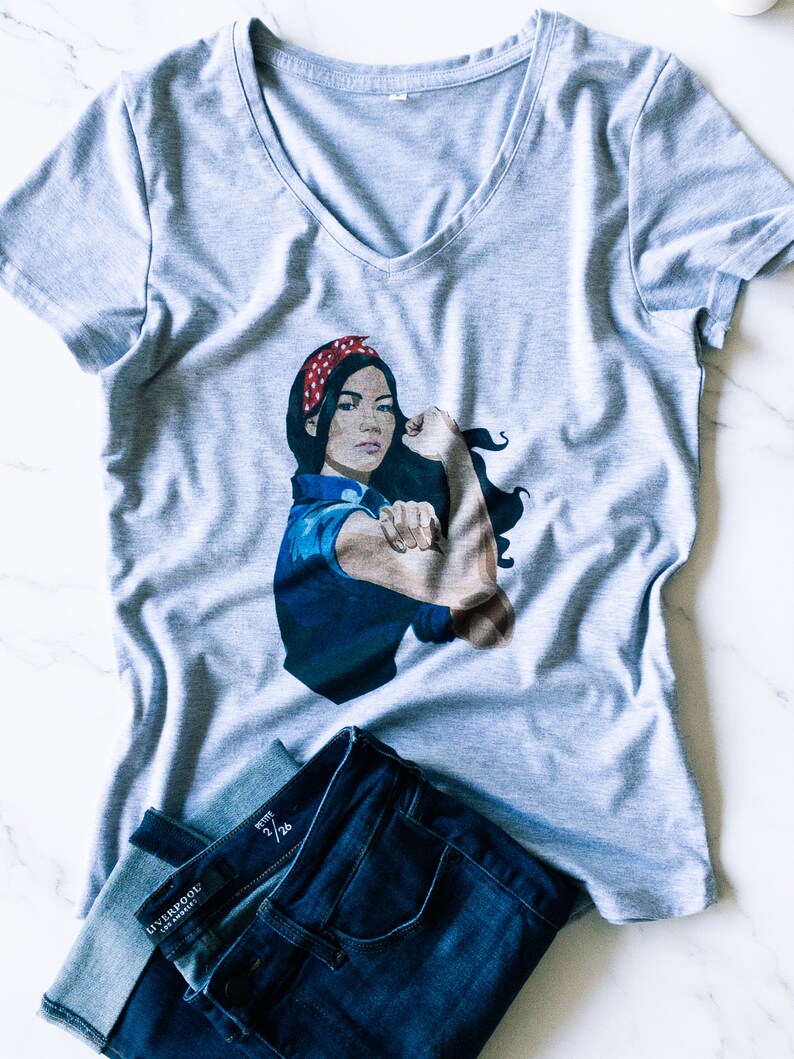 May include: A gray V-neck t-shirt with a graphic of a woman with a red bandana and a blue shirt, flexing her bicep. The woman is depicted in a classic "We Can Do It" pose. The t-shirt is paired with blue jeans.