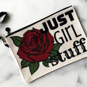 Peut inclure: Une pochette zippée en toile blanche avec une sangle noire. La pochette a une rose rouge avec des feuilles vertes et le texte "JUST GIRL STUFF" en noir.
