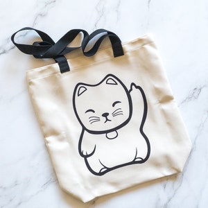 May include: A beige tote bag with black handles featuring a black and white illustration of a waving cat. The cat has a smiling face and a small collar. The bag is made of a canvas-like material.
