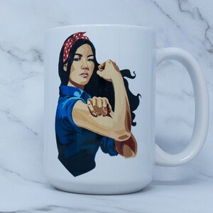 May include: White ceramic mug with a graphic of a woman with dark hair wearing a red and white polka dot headband, a blue shirt, and a flexed arm. The woman is depicted in a "We Can Do It!" style pose.