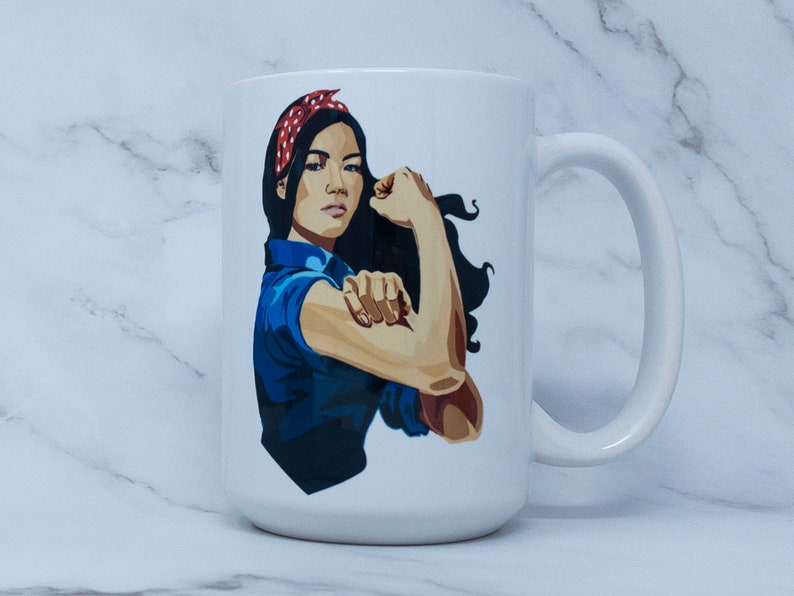 May include: White ceramic mug with a graphic of a woman with dark hair wearing a red and white polka dot headband, a blue shirt, and a flexed arm. The woman is depicted in a "We Can Do It!" style pose.