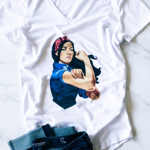 May include: White v-neck t-shirt with a graphic of a woman with a red headband and blue shirt, flexing her bicep. The woman is depicted in the "We Can Do It!" pose. The t-shirt is paired with a pair of dark blue jeans.