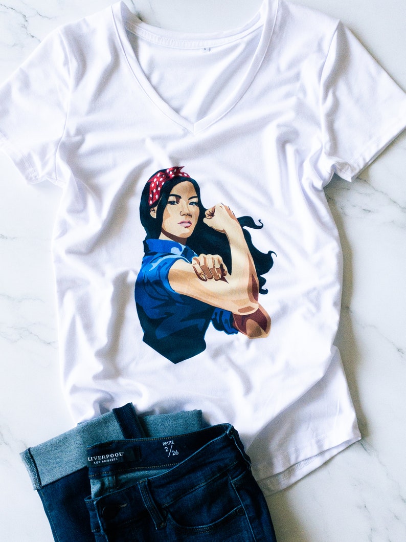 May include: White v-neck t-shirt with a graphic of a woman with a red headband and blue shirt, flexing her bicep. The woman is depicted in the "We Can Do It!" pose. The t-shirt is paired with a pair of dark blue jeans.