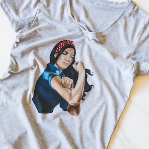 May include: A gray V-neck t-shirt with a graphic print of a woman with a red bandana, flexing her bicep. The woman is wearing a blue shirt and has dark hair.