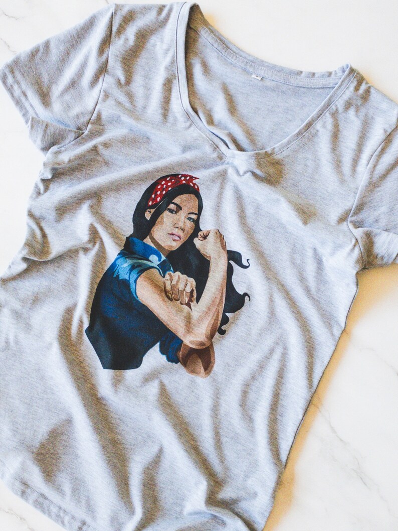 May include: A gray V-neck t-shirt with a graphic print of a woman with a red bandana, flexing her bicep. The woman is wearing a blue shirt and has dark hair.