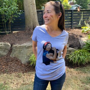 May include: A gray V-neck t-shirt with a graphic of a woman with a red bandana and blue shirt flexing her bicep. The graphic says "We Can Do It!"