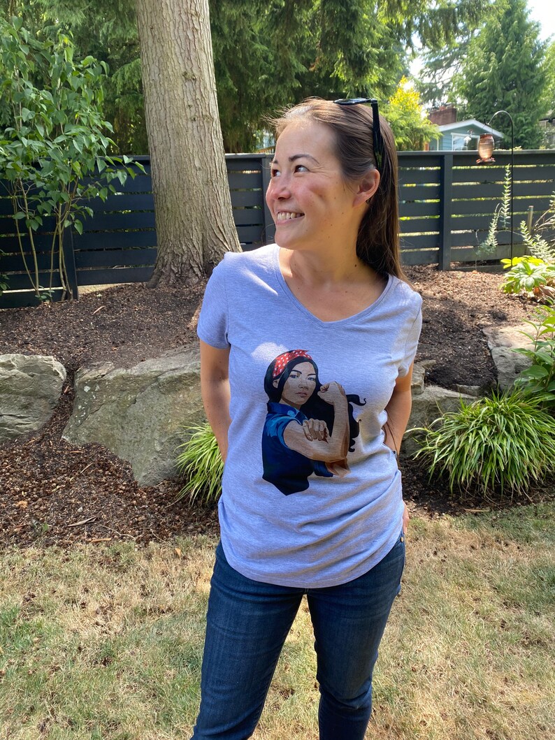 May include: A gray V-neck t-shirt with a graphic of a woman with a red bandana and blue shirt flexing her bicep. The graphic says "We Can Do It!"