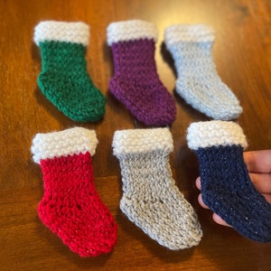 May include: Six miniature knitted Christmas stockings in various colors: green, purple, light blue, red, gray, and navy blue. Each stocking has a white cuff. One stocking is being held by a hand.