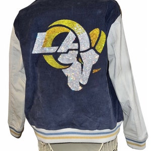 Puede incluir: Una chaqueta de béisbol azul y blanca con un gran logotipo de los Rams en strass en la espalda. El logotipo es de color dorado y plateado con un contorno blanco.