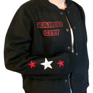 Puede incluir: Chaqueta de b&eacute;isbol negra con parches de estrellas rojas y blancas en las mangas. La chaqueta tiene un parche de "KANSAS CITY" con purpurina roja en la parte delantera.