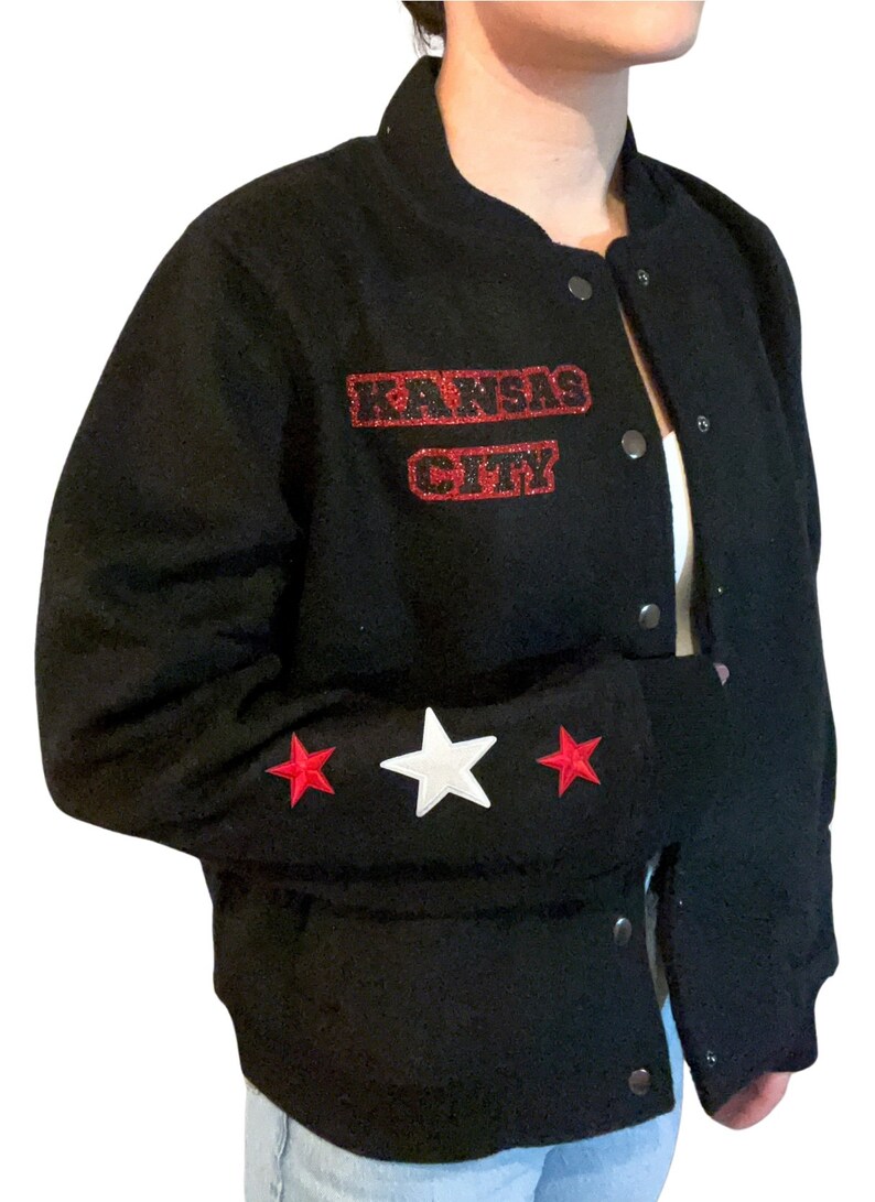 Puede incluir: Chaqueta de b&eacute;isbol negra con parches de estrellas rojas y blancas en las mangas. La chaqueta tiene un parche de "KANSAS CITY" con purpurina roja en la parte delantera.