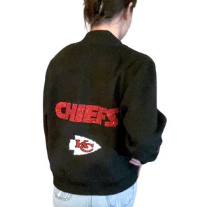 Puede incluir: Una chaqueta bomber negra con letras de purpurina roja que dice "CHIEFS" y una punta de flecha roja de purpurina con "KC" en su interior.