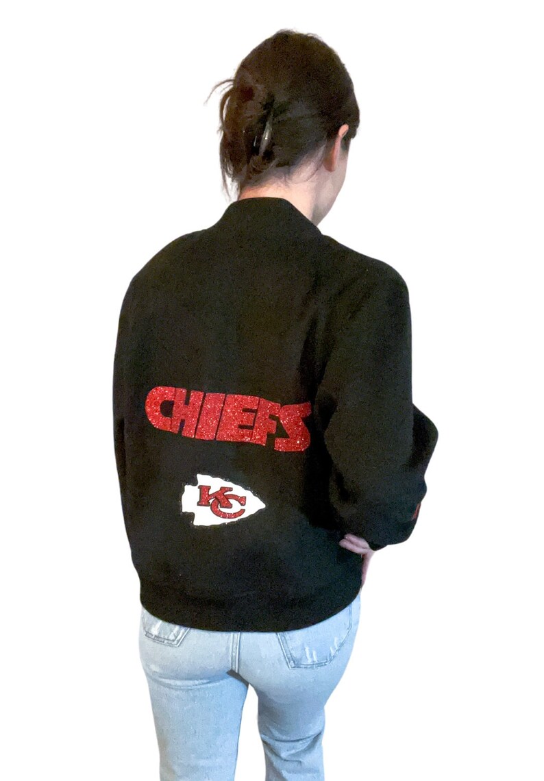 Puede incluir: Una chaqueta bomber negra con letras de purpurina roja que dice "CHIEFS" y una punta de flecha roja de purpurina con "KC" en su interior.