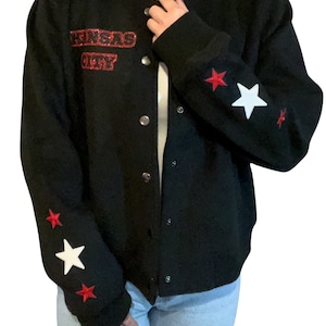 Puede incluir: Chaqueta bomber negra con parches de estrellas rojas y blancas en las mangas. La chaqueta tiene un parche rojo "Kansas City" en la parte delantera.